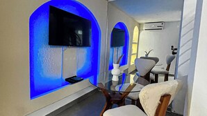 A modern interior with a flat-screen TV mounted on a wall with blue lighting, a dining area with a table and chairs, and an air conditioning unit on the wall. - Degn'An Holding Residence. (Abidjan)