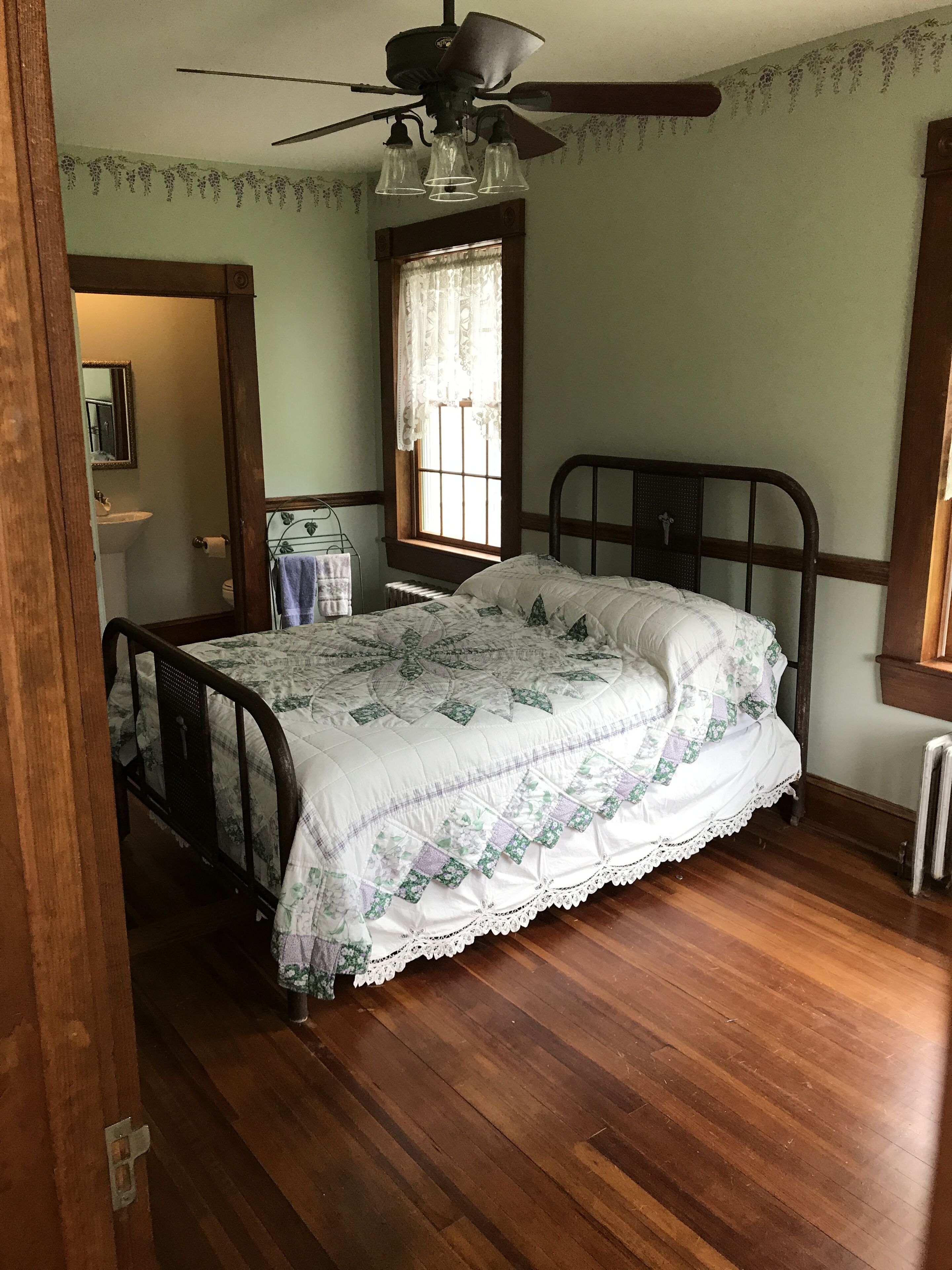 A bedroom with a wooden floor, a bed with a floral quilt, a ceiling fan, a window with lace curtains, and a radiator.
