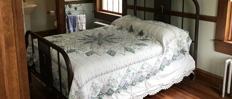 A bedroom with a wooden floor, a bed with a floral quilt, a ceiling fan, a window with lace curtains, and a radiator.