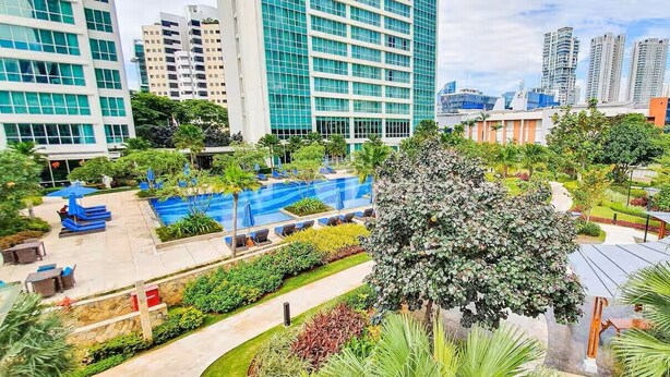 Un hotel moderno con piscina en la azotea, exuberante vegetación y edificios altos al fondo.