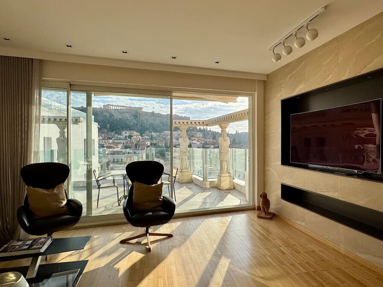 Une pièce spacieuse avec un grand balcon, un téléviseur fixé au mur et deux fauteuils modernes.