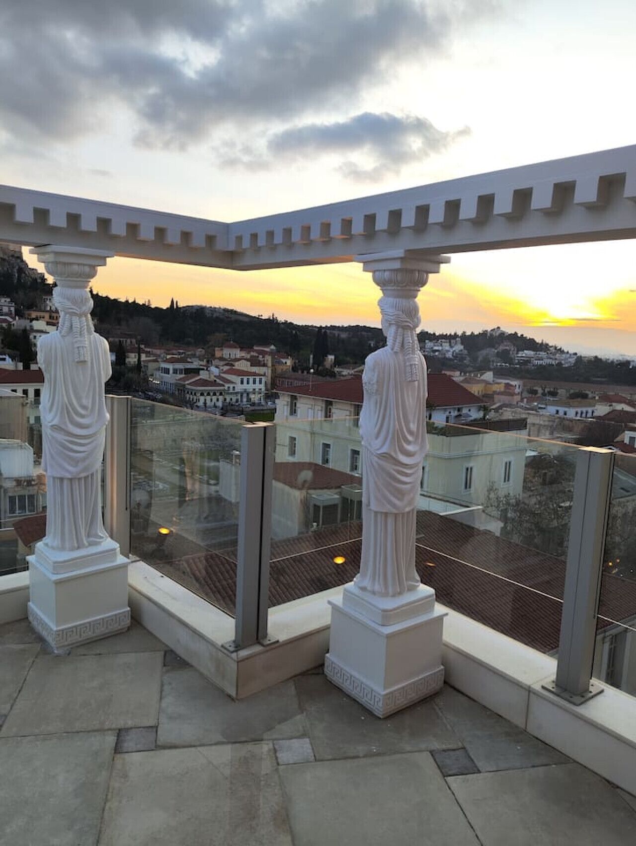 Un balcon doté de colonnes blanches et d’une balustrade en verre, offrant une vue sur le paysage urbain au coucher du soleil.