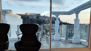 A modern room with a large window overlooking a cityscape and a view of ancient ruins. - Luxury Acropolis-View Apartment with Panoramic Terrace | Monastiraki (Athina)
