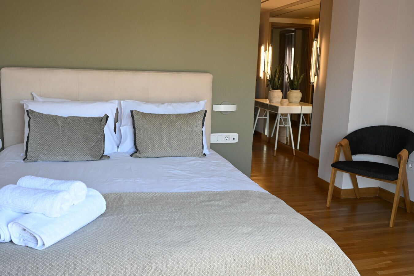A neatly made bed with a grey duvet and two pillows, a wooden chair, and a small desk with a chair.