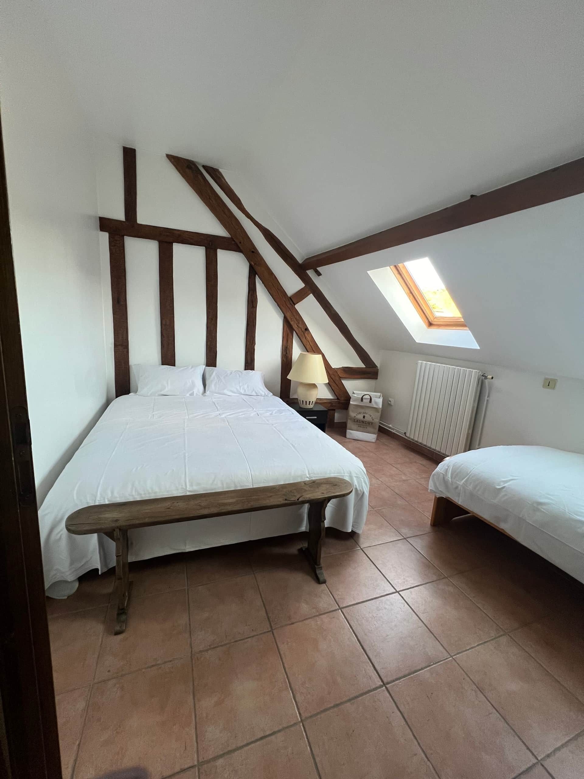 Ein Schlafzimmer mit einem großen Bett, einem kleineren Bett, einer Holzbank, einem Oberlicht und einer Heizung.