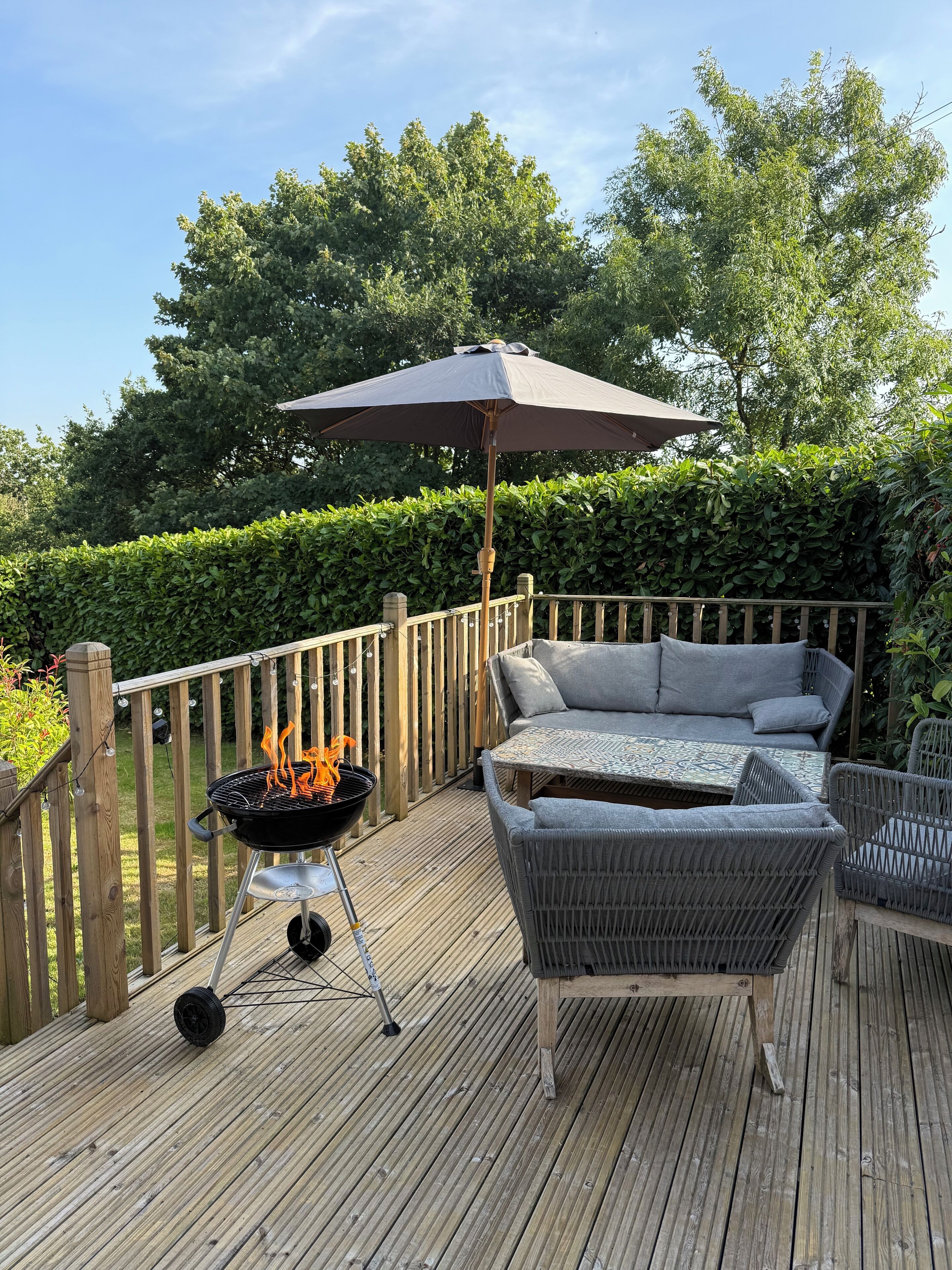 Eine Holzterrasse mit Grill, Gartenmöbeln und einer Hecke im Hintergrund.