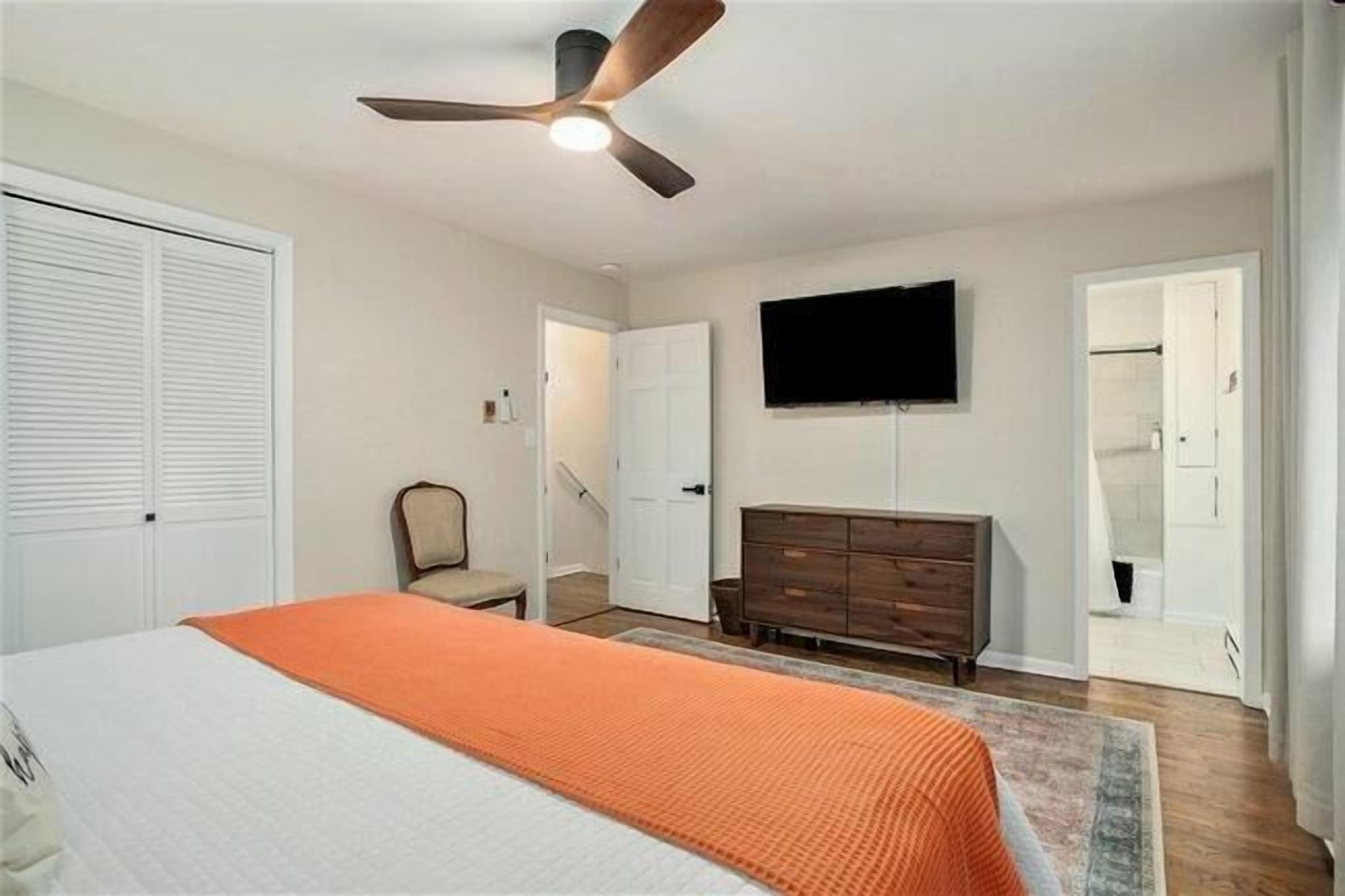 A bedroom with a bed, a ceiling fan, a TV mounted on the wall, a dresser, and a walk-in closet.