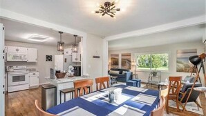 Una cocina moderna con armarios blancos, un comedor con mantel azul y un amplio ventanal con vista a zonas verdes.