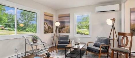 Een moderne woonkamer met een bank, fauteuils, een salontafel en een lamp. Grote ramen bieden uitzicht op groen buiten.