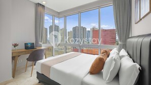 Une chambre d’hôtel avec une grande fenêtre, un lit avec des oreillers blancs et bruns, un bureau avec un ordinateur portable et une chaise.