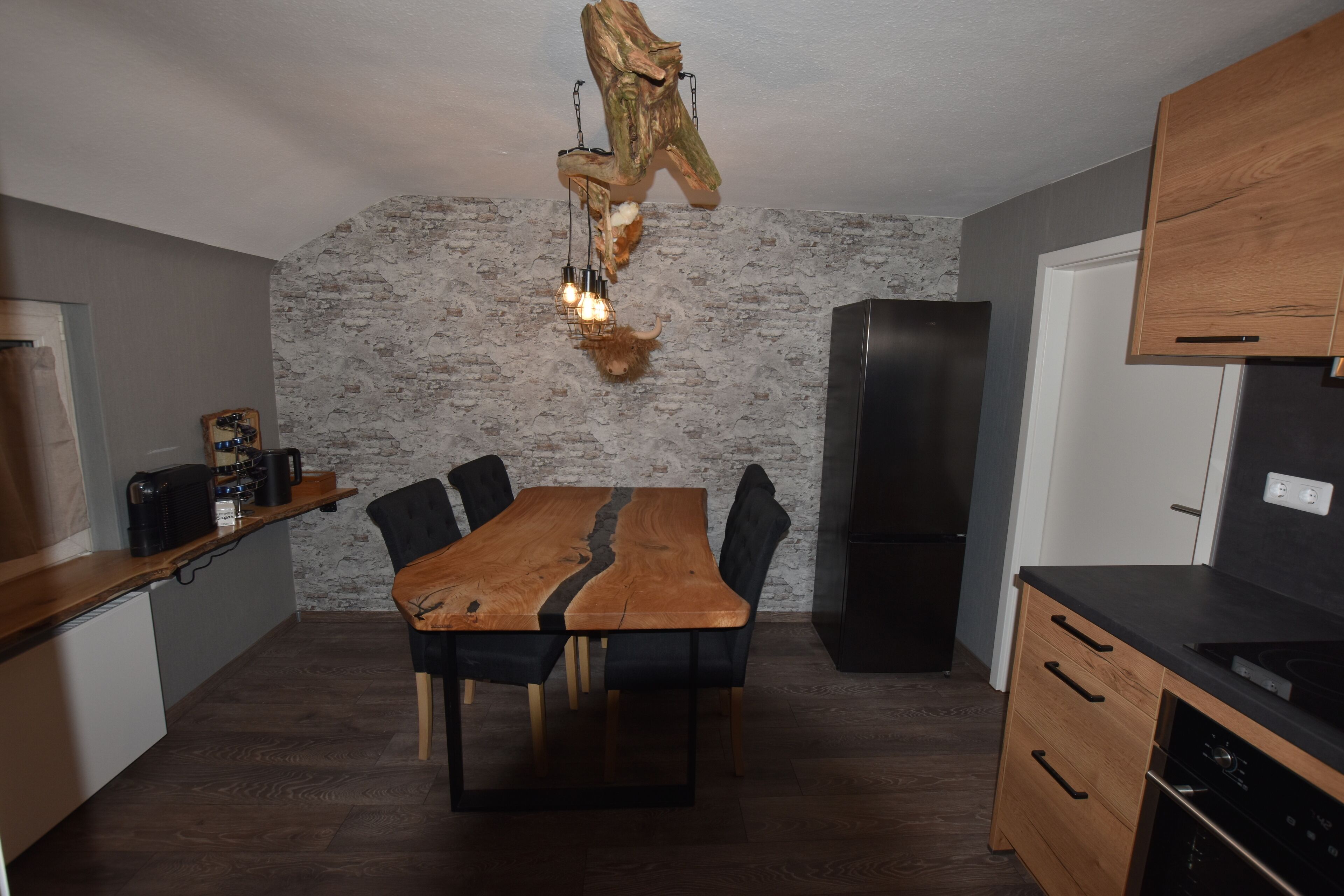 A modern kitchen with a wooden dining table, black chairs, a stone wall, and a hanging light fixture.