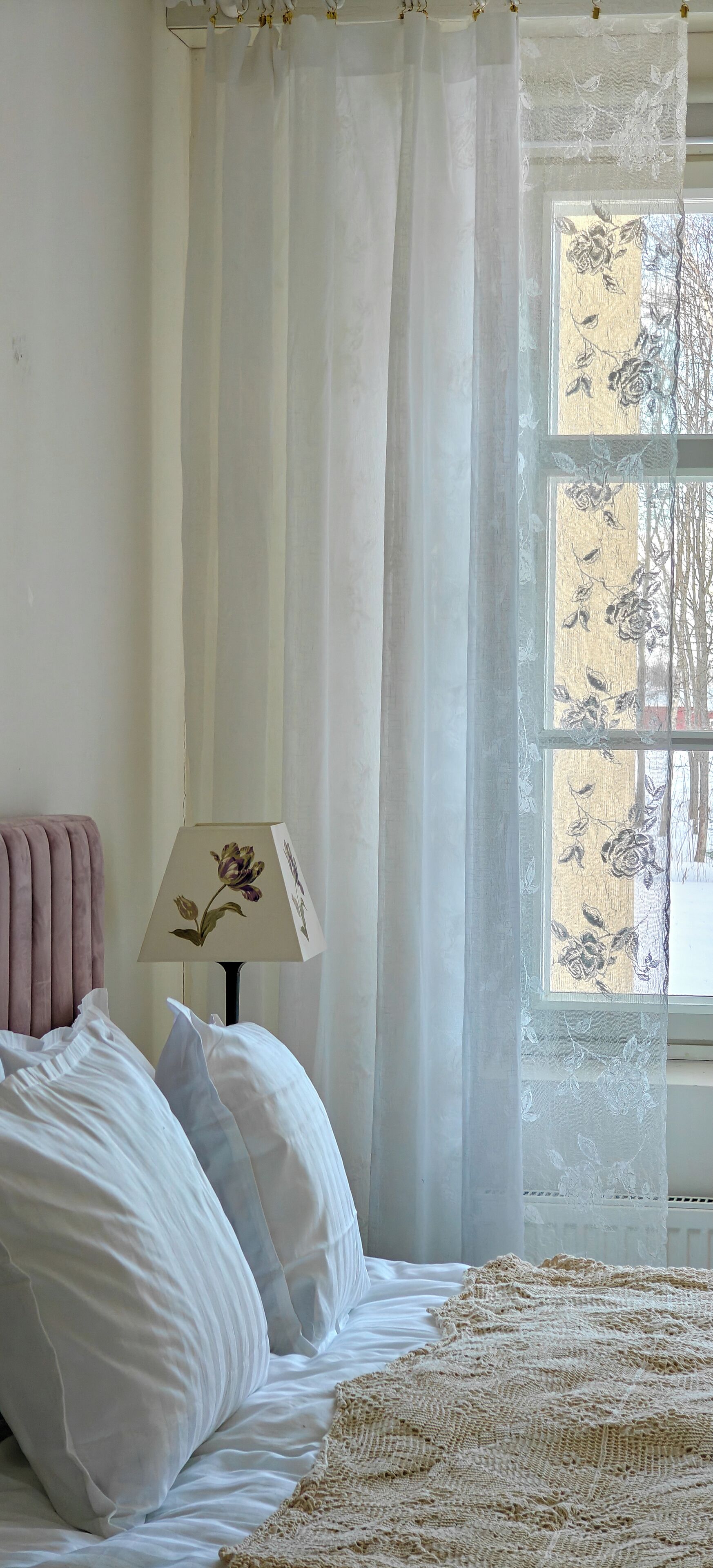 A bedroom with a bed, white pillows, a knitted blanket, a pink headboard, a lamp with a floral design, sheer curtains, and a window with a floral pattern.
