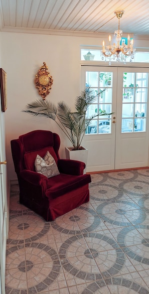 Una habitación amplia con un sillón tapizado en terciopelo rojo, una planta grande en maceta, un reloj de pared decorativo y una araña.