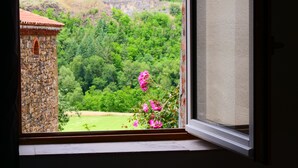 A window view of a stone building with a red roof, surrounded by lush greenery and hills. - House "Les sauvages" - Nature and charm on the banks of the Allier 8 to 10 people (Blassac)