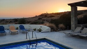 Un espace piscine avec des chaises longues et une vue sur le coucher de soleil, qui se reflète sur un paysage vallonné.