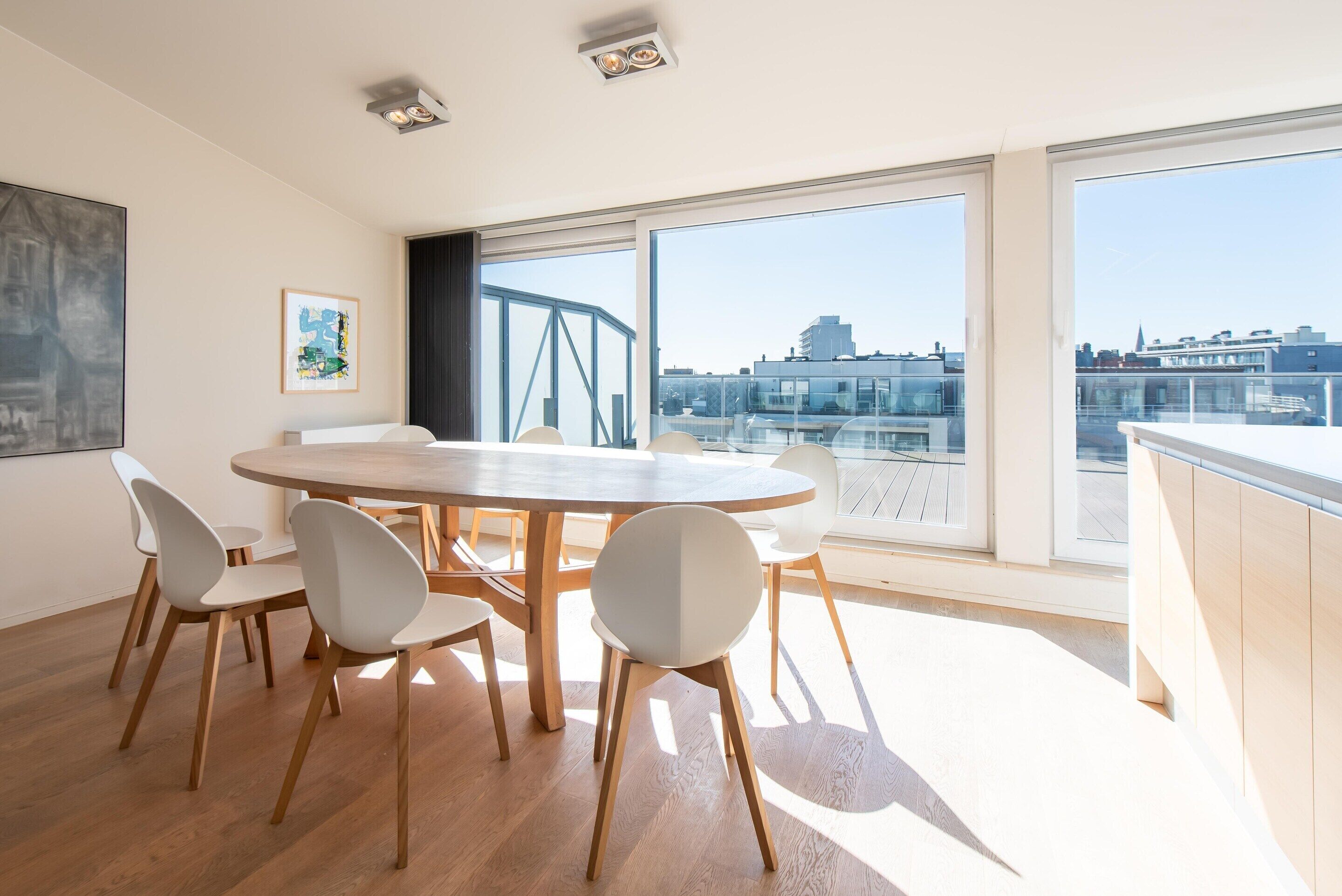 A modern dining area with a round table and six chairs, large windows with a city view, and a painting on the wall.