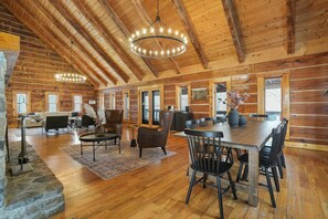 A spacious log cabin interior with a high ceiling, wooden beams, a dining area with a large table and chairs, a fireplace, and a stone accent wall. - Lodge at Wildcat Hollow | Game Room, Hot Tub (Rockbridge)