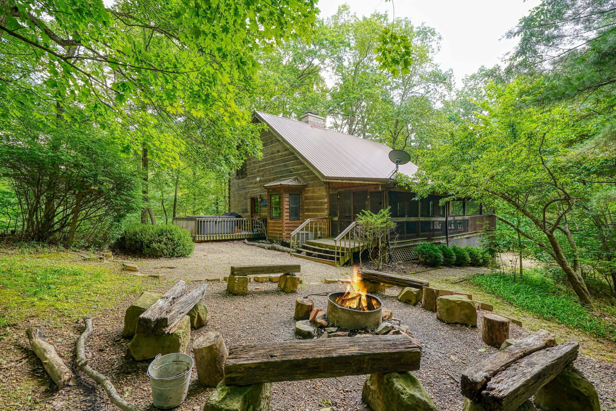 A rustic cabin with a fire pit in the center, surrounded by wooden benches and a gravel area.