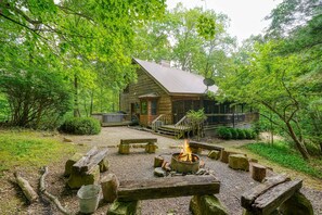 A rustic cabin with a fire pit in the center, surrounded by wooden benches and a gravel area.