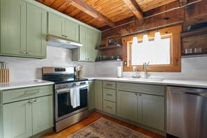 A kitchen with green cabinets, stainless steel appliances, a window with blinds, and a wooden ceiling.
