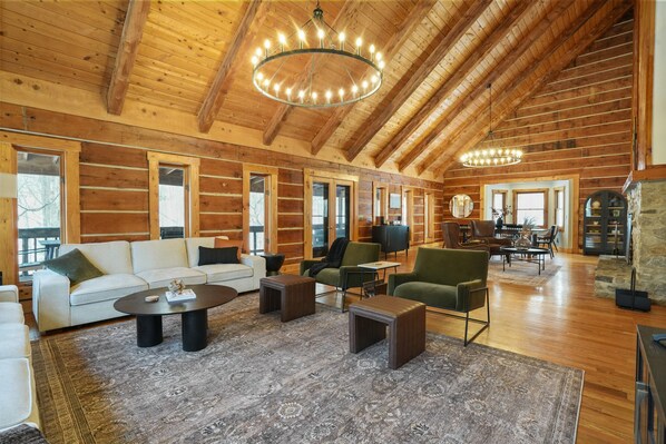 A spacious log cabin interior with a high ceiling, wooden beams, and a large chandelier. There are multiple seating areas with sofas and chairs, and a stone fireplace.