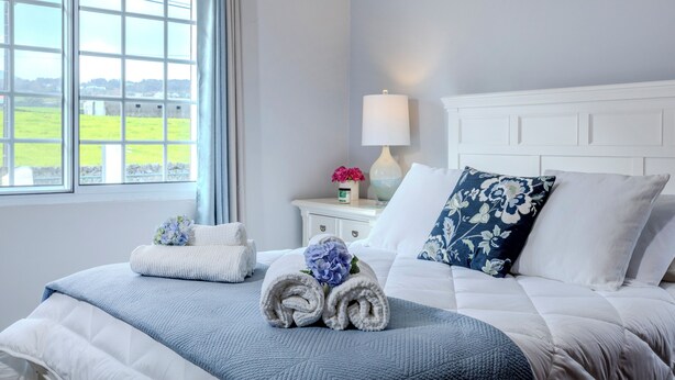 A neatly made bed with rolled towels and a flower, a bedside table with a lamp and a vase of flowers, and a window with a view of a green landscape.