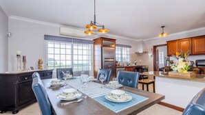 A dining area with a wooden table set for four, surrounded by blue chairs. There is a kitchen in the background with wooden cabinets and a stainless steel refrigerator.