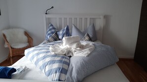 Una cama bien hecha con ropa de cama a rayas y varios almohadones, una silla blanca con un cojín mullido, y un cabecero blanco.
