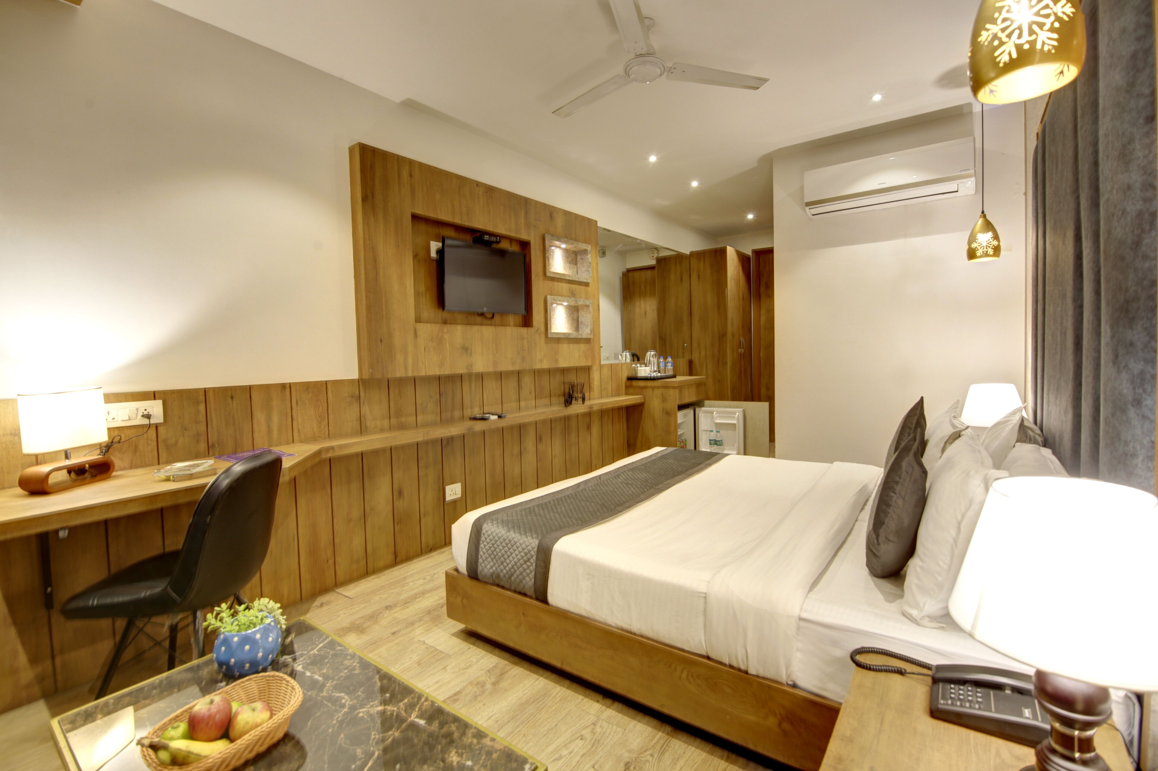 Ett hotellrum med en väggpanel i trä, en säng med kuddar, ett skrivbord med en stol, en telefon och en tv som är monterad på väggen.