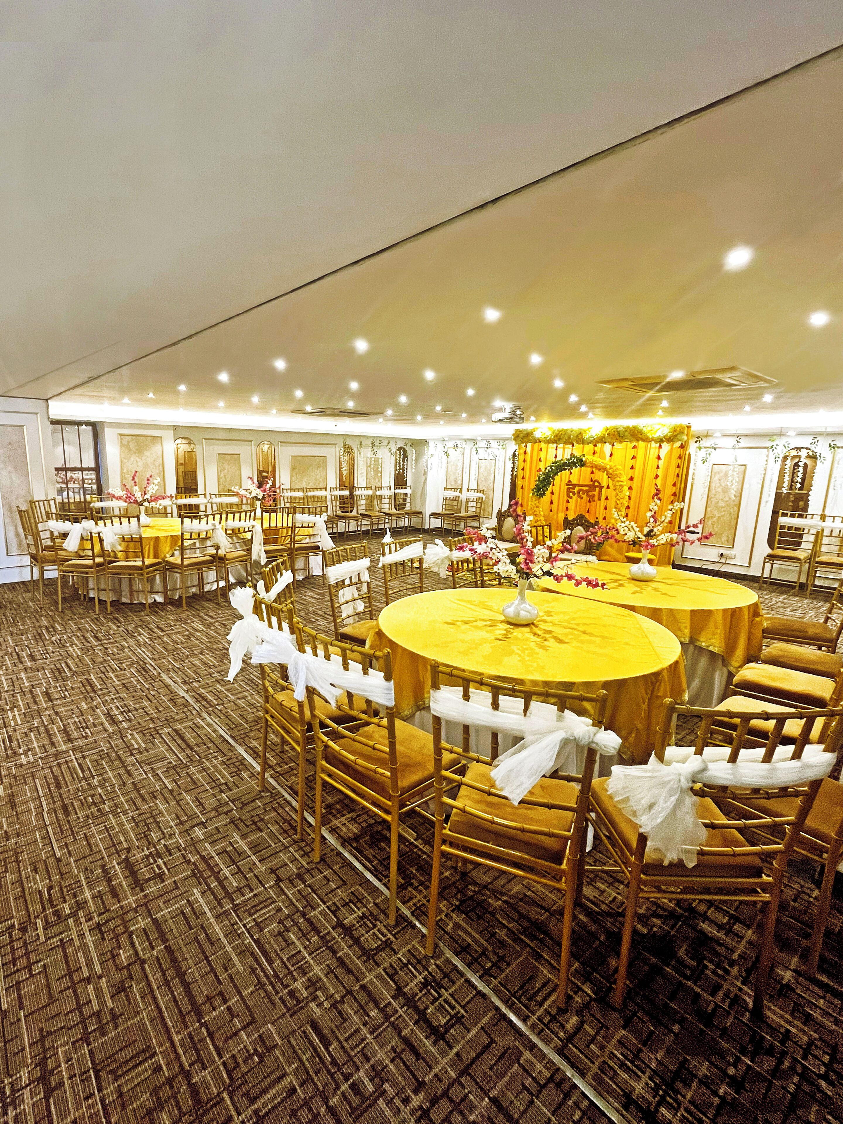 A banquet hall with round tables and chairs, a carpeted floor, and a stage with floral decorations.