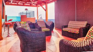 Un espacio de estar al aire libre con techado, muebles de mimbre y cojines de colores, con vistas a una piscina y al mar.