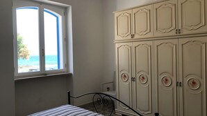 A bedroom with a bed, a window with a view of the sea, and a white wardrobe with decorative handles.