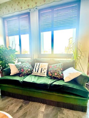 Un divano verde con cuscini floreali e un cuscino con la scritta “HOME SWEET HOME”, posizionato davanti a una finestra con persiane.