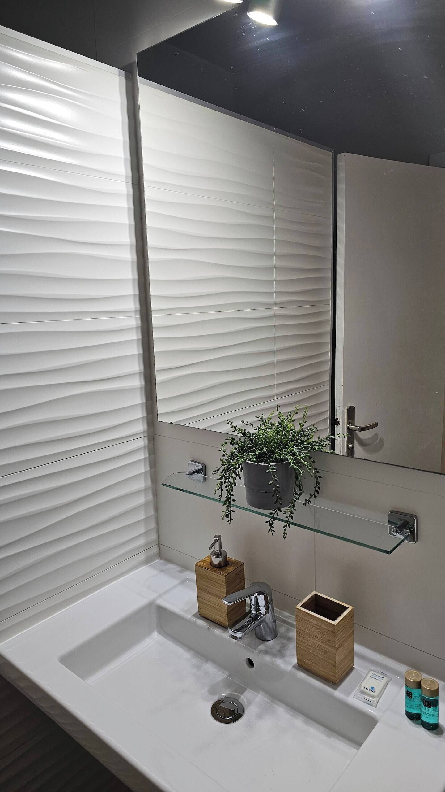 A modern bathroom with a white sink, a mirror, a potted plant, and a glass shelf.
