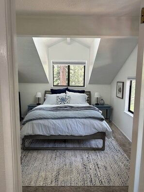 A bedroom with a sloped ceiling, a bed with striped bedding, two bedside tables with lamps, and a window with a view of trees. - Quiet 1 Bdrm walk to beach & trails. Close to ski areas. (Kings Beach)