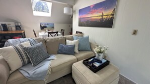 A living room with a beige sofa, a coffee table with a vase of flowers, a painting of a sunset, and a dining area with a table set for two. - Quiet 1 Bdrm walk to beach & trails.  Close to ski areas. (Kings Beach)