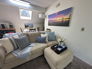 A living room with a beige sofa, a coffee table with a vase of flowers, a painting of a sunset, and a dining area with a table set for two. - Quiet 1 Bdrm walk to beach & trails. Close to ski areas. (Kings Beach)