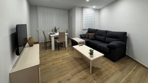 Una sala de estar moderna con un sofá oscuro, una mesa de centro blanca, un televisor y un comedor con una mesa y sillas.