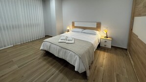 Un dormitorio moderno con piso de madera, una cama grande con ropa blanca, dos lámparas de noche y un ventanal con persianas.