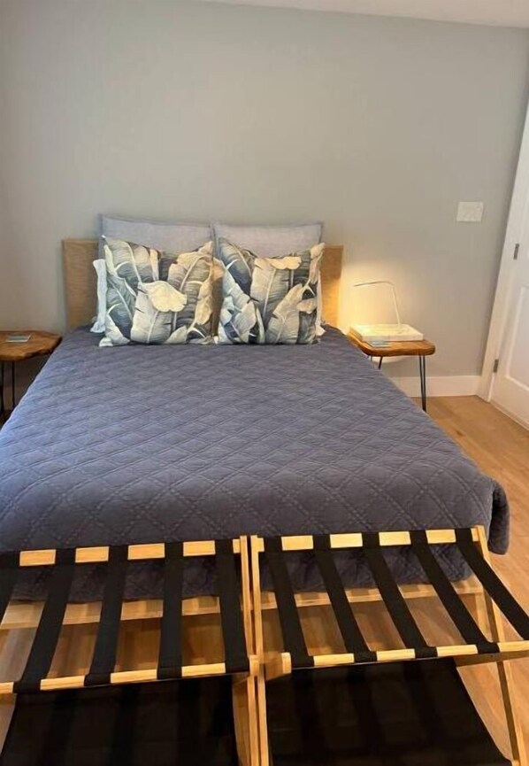 Una cama con cubrecolcha gris acolchado, dos almohadas con estampado de hojas y un cabecero de madera.