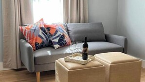 A modern living room with a grey sofa, colorful pillows, a small wooden coffee table with a wine bottle and glasses, and a beige ottoman. - Coastal Private Guest Suite (Cohasset)