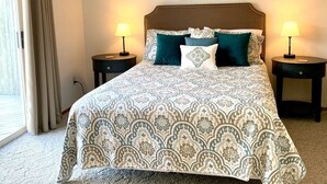 Una cama bien hecha con ropa de cama estampada, dos mesitas de noche con lámparas, un cuadro en la pared y una ventana con cortinas.