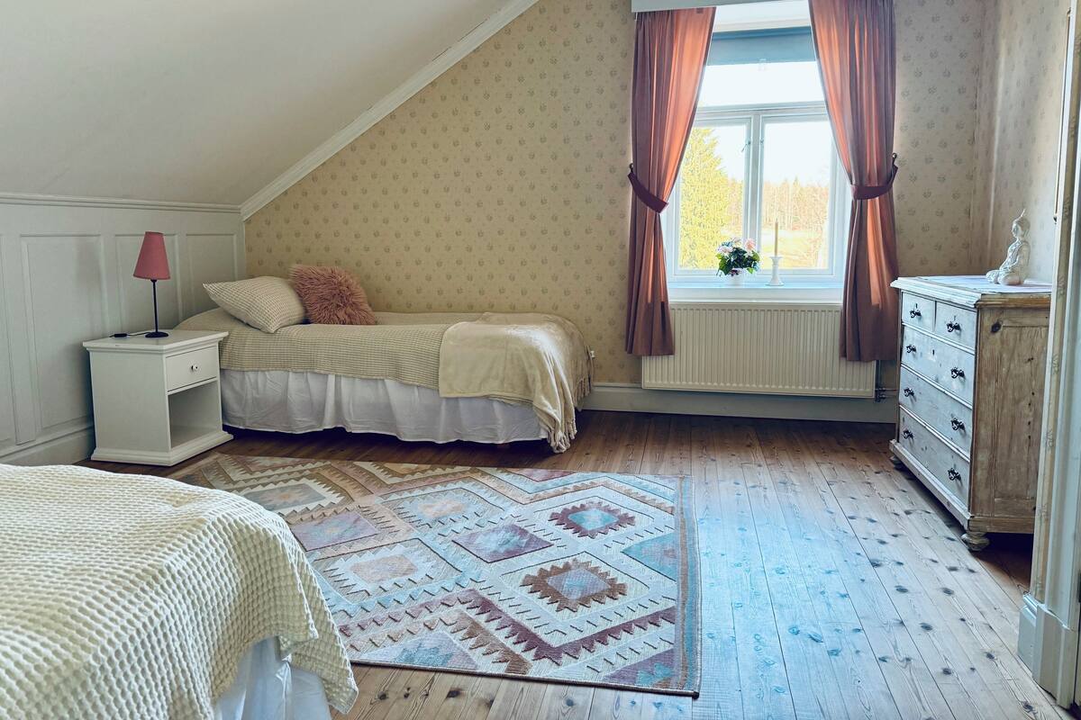 Un dormitorio con techo inclinado, una cama con colcha estampada, una mesita de noche con lámpara, un aparador y una ventana con cortinas.