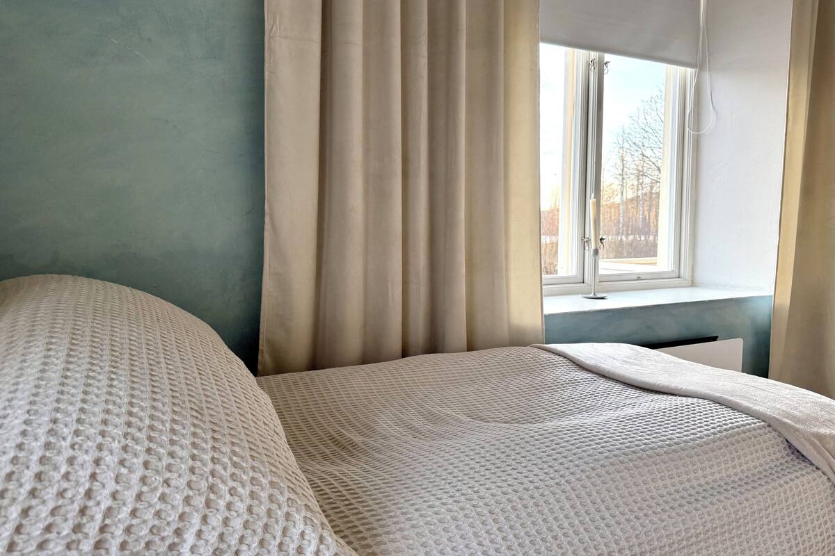 Un dormitorio con una cama, cortinas color beige y una ventana con vistas a árboles.