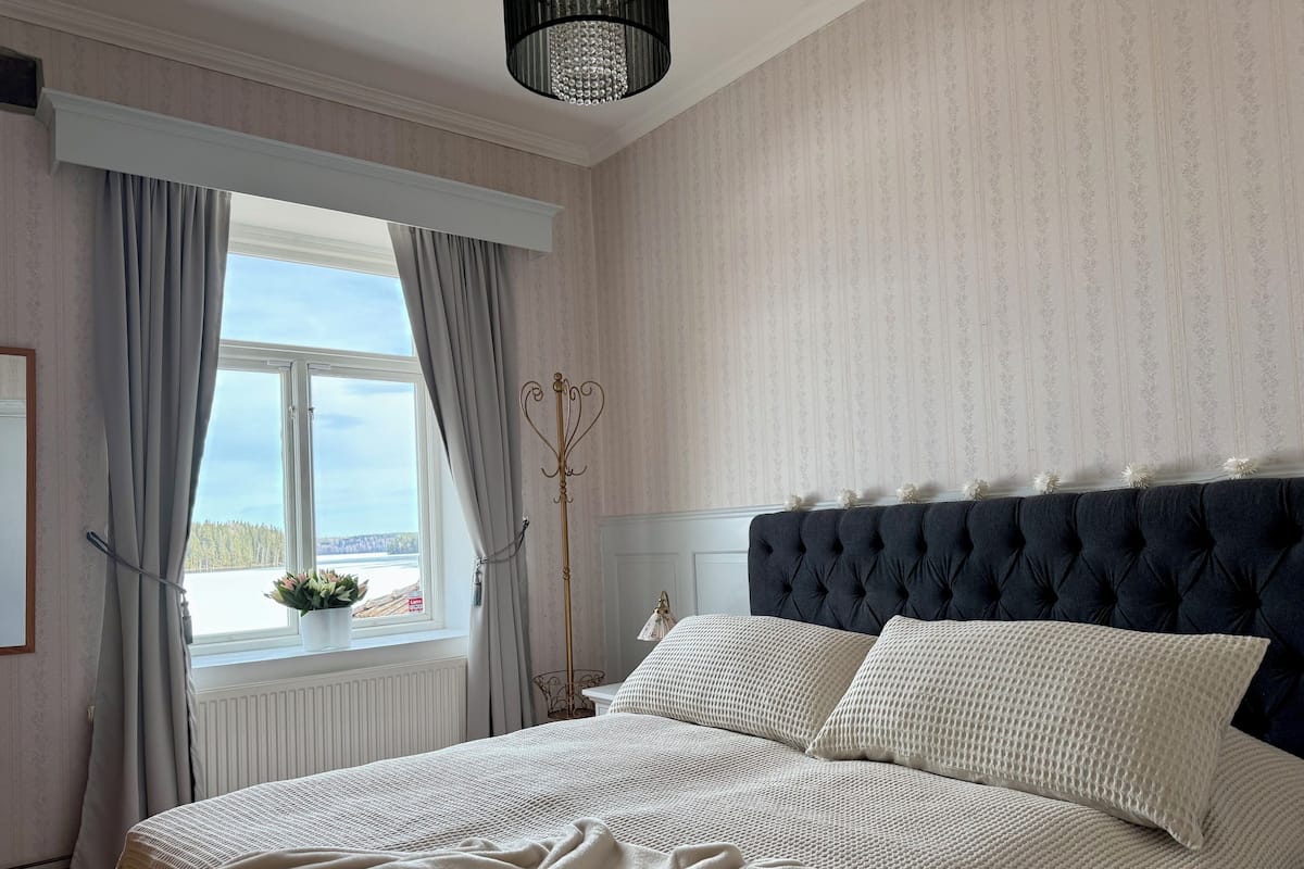 Un dormitorio con una cama grande, una ventana con cortinas, una araña y vistas al exterior.