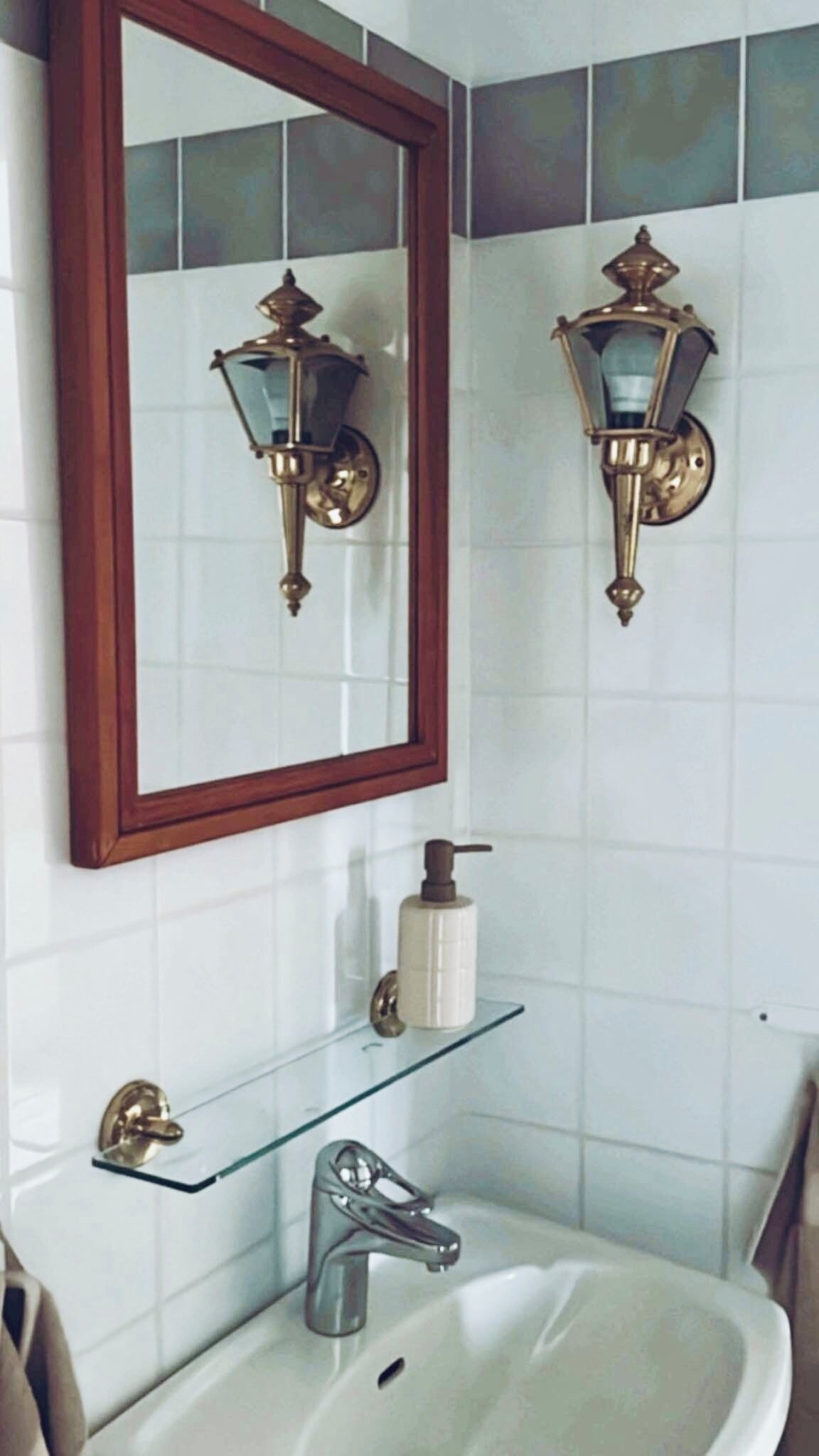 A bathroom with a white sink, a mirror with a wooden frame, two wall-mounted light fixtures, and a glass shelf with toiletries.