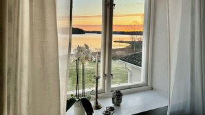 Une fenêtre donnant sur un lac, avec une vue sur un coucher de soleil, une statue de Bouddha et un vase rempli de fleurs posé sur le rebord de la fenêtre.