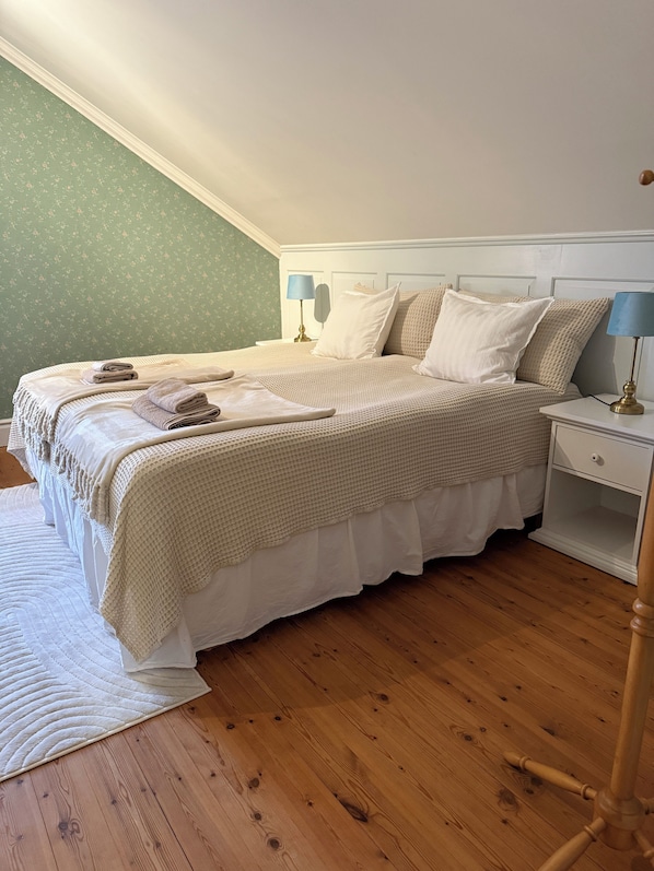 Una cama bien hecha con una colcha blanca y beige, dos lámparas de noche y suelo de madera.