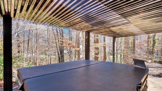 An outdoor seating area with a table and chairs, surrounded by trees and a metal roof structure.