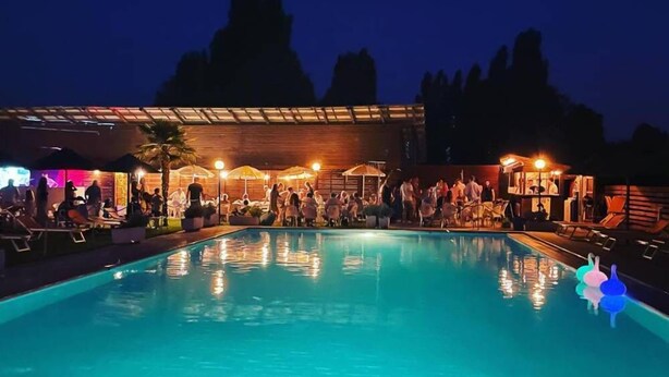 A swimming pool with lounge chairs and umbrellas, surrounded by a building with a wooden facade and a covered area with people. - TENUTA SANTO STEFANO - HOTEL (Campagnola Emilia)